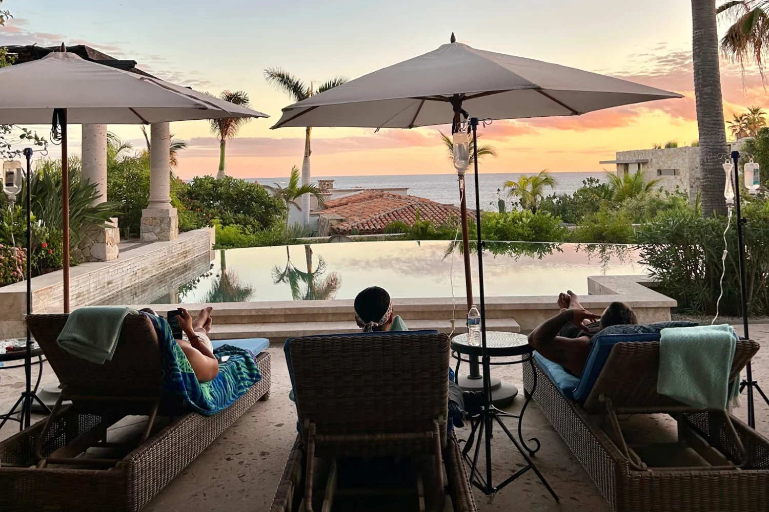A group receiving IV therapy together on poolside loungers at a private villa in Los Cabos, with IV stands beside each guest and the ocean at sunset beyond the infinity pool